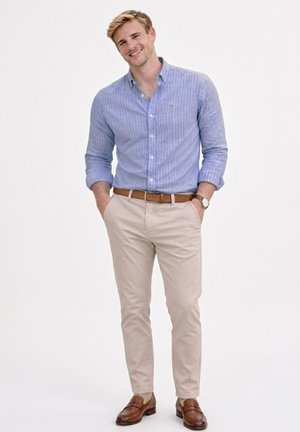 Joven sonriente, vistiendo una camisa de botones a rayas azules, pantalones beige, cinturón marrón, mocasines marrones y un reloj de pulsera, de pie con las manos en los bolsillos.