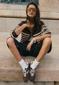 Brązowo-biały kardigan w paski, czarne krótkie spodenki ze sztucznej skóry, białe skarpetki i brązowe trampki z kremowymi akcentami. Okulary z żółtą soczewką, siedząca poza.