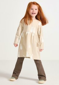 Beige pitkähihaine mekko, jossa on kerrostettu vyötärö, esittelee valkoisia ja ruskeita pupumalleja, yhdistettynä ruskeisiin corduroy-housuihin ja vihreisiin kenkiin.