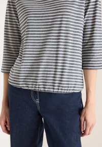 Femme portant un jean bleu marine et un haut à manches trois-quarts à rayures horizontales grises et blanches, debout les mains détendues le long du corps.