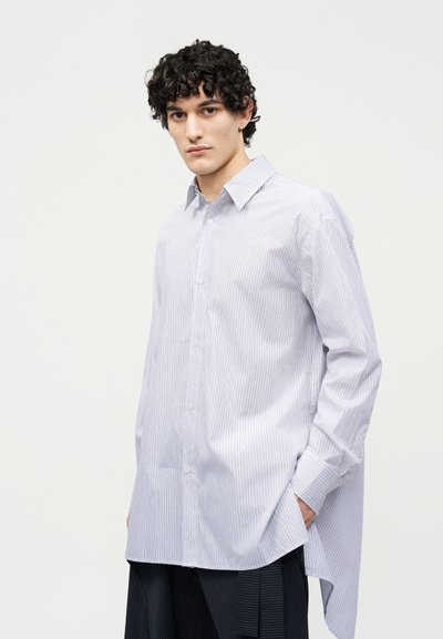 Junger Mann mit lockigem Haar, der ein locker sitzendes, weißes Langarmhemd mit dünnen vertikalen Streifen und dunkel gestreifte Anzughose trägt, steht mit den Händen in den Taschen.