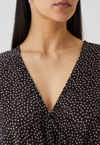 Kvinna med rakt mörkt hår, bärande pärlörhängen och en svart klänning med beiga prickar och en V-formad halsringning.