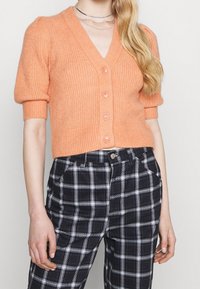 Orange stickad cardigan med korta ärmar, v-ringning och knäppning. Kombinerad med svart och vit rutig hög midja byxor.