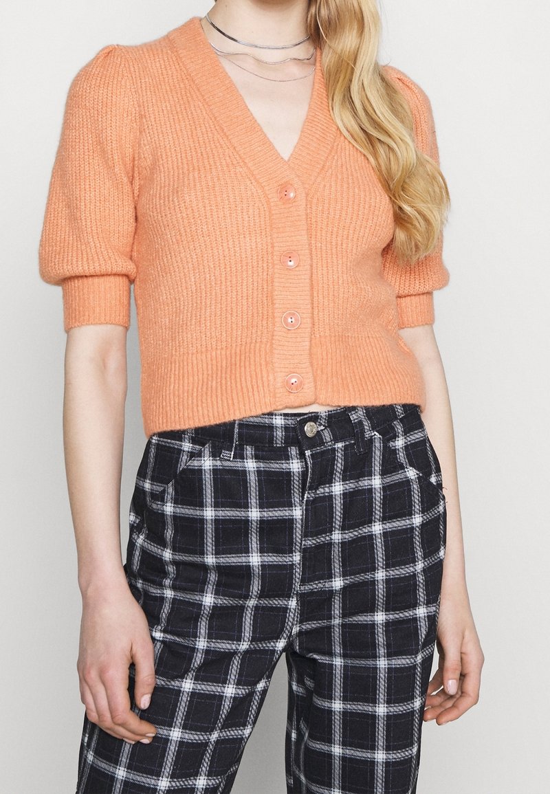 Orange stickad cardigan med korta ärmar, v-ringning och knäppning. Kombinerad med svart och vit rutig hög midja byxor.
