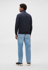 Felpa navy con colletto alto, polsini a costine e vestibilità ampia, abbinata a jeans azzurri chiari, larghi e sneaker nere.