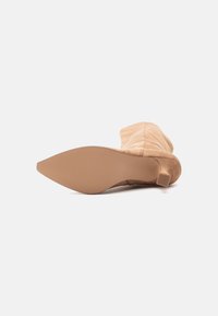 Stivale beige in suede a punta, alto fino al polpaccio, sdraiato su un lato che mostra la suola liscia e il piccolo tacco a blocco.