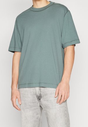 Mann trägt ein locker sitzendes, kurzärmliges T-Shirt in Salbeigrün, kombiniert mit hellgrauen, gewaschenen Jeans vor einem einfarbigen Hintergrund.