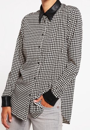 Černobílá košile s knoflíky v pepito vzoru s černým koženým límcem a manžetami, nošená s světle šedými džínami, ruka v kapse.