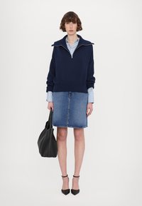 Pulover tricotat navy cu guler înalt și fermoar pe jumătate, purtat deasupra unei cămăși în dungi albastre deschise și a unei fuste din denim. Geantă neagră și tocuri.