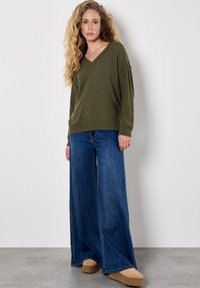 Pull en v olive avec ourlet côtelé, associé à un jean en denim à jambe large de couleur bleu foncé. Le mannequin porte des chaussures à plateforme beiges, se tenant devant un fond clair.