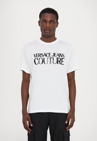 Biała bawełniana koszulka z nadrukiem "VERSACE JEANS COUTURE" w wyrazistych czarnych literach na klatce piersiowej. Okrągły dekolt i krótkie rękawy.