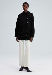 Vrouw in een oversized zwart knopentrui, witte wijde broek en zwarte spitse haklaarzen, staande op een eenvoudige lichte achtergrond.
