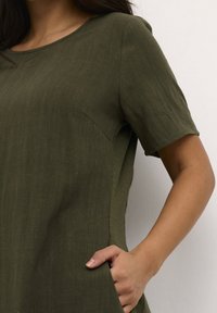 Vestido corto de manga corta en verde oliva, confeccionado en una tela texturizada y ligera, con escote redondeado y un detalle de bolsillo lateral.