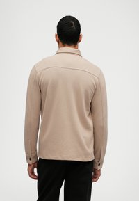 Camicia beige a maniche lunghe con colletto, presenta dettagli di cucitura frontale, polsini con bottoni e un tessuto testurizzato. Indossata con pantaloni neri.