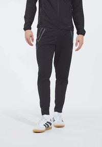 Veste de sport noire et pantalon en matériau léger, avec des poches zippées et un design ajusté. Baskets blanches avec des rayures noires.