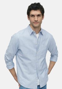Chemise bleu clair à boutons, à manches longues, avec une poche poitrine et un tissu légèrement texturé, superposée à un t-shirt blanc.