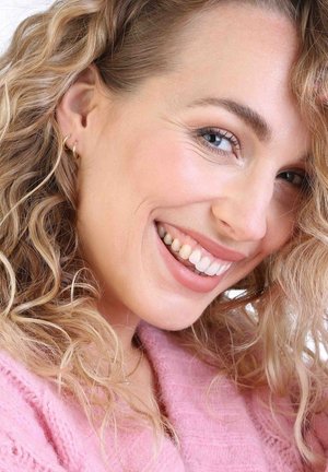 Gros plan sur une personne souriante avec de longs cheveux bouclés châtain clair. Elle porte des boucles d'oreilles en or et un pull en tricot rose doux. Peau claire et dents blanches.