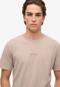 Lys beige bomulds T-shirt med korte ærmer og rund halsudskæring. Har et lille broderet "SUPERDRY" logo på brystet.