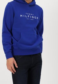 Felpa blu in cotone con tasca frontale e testo del logo. Presenta un cappuccio regolabile con lacci e polsini a costine, abbinata a jeans scuri.