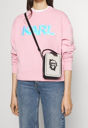 Černá a béžová kabelka přes rameno s prošitým okrajem a grafikou lva; nošená s růžovou mikinou a modrými džíny.