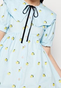 Jasnoniebieska sukienka z dużym, falbaniastym kołnierzem, czarnym krawatem i żółtymi haftowanymi cytrynami, z perłowymi guzikami z przodu.