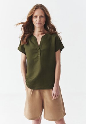 Camicia in lino verde oliva con scollo a V e maniche corte, dotata di una chiusura a bottoni. Abbinata a pantaloni sartoriali chiari in tessuto simile.