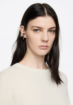 Pendientes de oro y plata con un acento de gema clara, llevados en la oreja izquierda. El sujeto lleva un suéter de punto color crema claro.