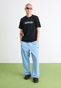 T-shirt preta com o logotipo azul "ADIDAS", calças de moletom azul claro com riscas pretas, e tamancos pretos. Tecido suave, ajuste descontraído.