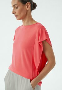 Koralowy top z krótkim rękawem, okrągłym dekoltem i luźnym krojem. Wykonany z miękkiej tkaniny, z delikatnym fałdowaniem i lekką konstrukcją.