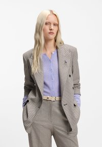 Blazer croisé en pied-de-poule marron et crème, associé à un chemisier boutonné violet clair et à une ceinture crème. Coupe ajustée.