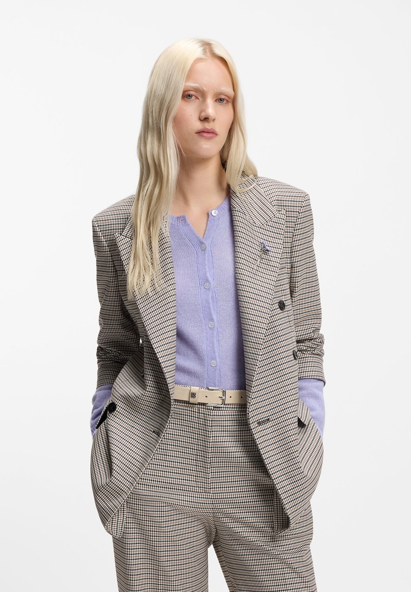 Blazer croisé en pied-de-poule marron et crème, associé à un chemisier boutonné violet clair et à une ceinture crème. Coupe ajustée.
