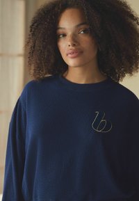 Donkerblauwe textuur sweatshirt met een ronde hals, voorzien van een gouden geborduurd logo op de linkerkant van de borst. Lange mouwen.