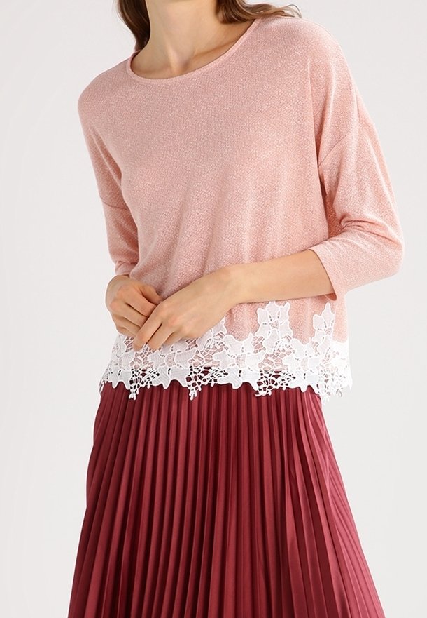 Femme portant un haut rose clair à manches trois-quarts avec un ourlet en dentelle blanche et une jupe plissée bordeaux taille haute, les mains jointes.