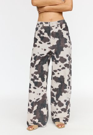 Pantalon à jambes larges avec motif imprimé vache noir et blanc porté avec des sandales beiges à bout ouvert, personne debout bras croisés au-dessus de la taille.