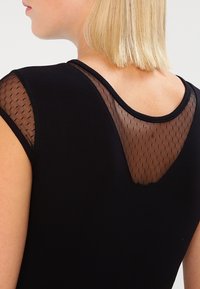 Top negro con un yugo de malla transparente con un patrón de diamantes, mangas cortas y escote redondeado. Tela suave y diseño ajustado.