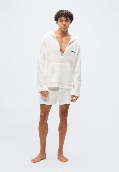 Jeune homme debout pieds nus portant un pull à capuche blanc à manches longues et un short assorti avec une ouverture en V profonde.