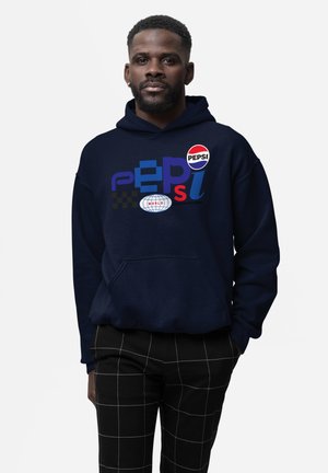 Uomo che indossa una felpa blu navy con loghi colorati della Pepsi e pantaloni neri con motivo a griglia bianca, in piedi su uno sfondo semplice.