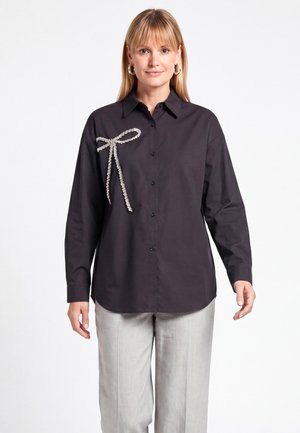 Donna che indossa una camicia nera a maniche lunghe con bottoni e un fiocco decorativo di perle sul lato sinistro e pantaloni grigio chiaro, in piedi su sfondo bianco.