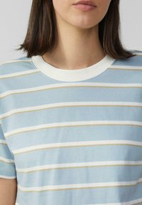 Vrouw in een lichtblauw, witte en beige horizontaal gestreepte T-shirt met een witte ronde halslijn, gezien van de nek tot halverwege de torso.