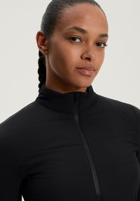 Donna con capelli intrecciati che indossa una giacca nera con zip, rivolta leggermente a sinistra, espressione neutra, su uno sfondo chiaro e semplice.