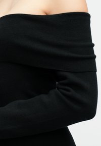 Maglione nero scivolato sulle spalle con texture a coste, caratterizzato da maniche lunghe e una vestibilità aderente. Il materiale sembra morbido ed elastico.