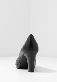 Černá kožená boty na vysokém podpatku pohledem zezadu, s kulatým podpatkem a hladkým povrchem na bílé ploše.