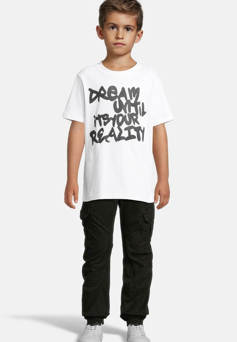 Balta medvilninė marškinėliai su juodu graffiti stiliaus tekstu, rašančiu "SVAJOK, KOL TAPSI TAI REALYBE." Suderinti su juodomis cargo kelnėmis, elastinėmis rankovėmis ir baltais sportiniais bateliais.