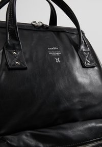 Bolso de mano de cuero negro con forma redondeada, doble asa, herrajes plateados y un logo en la parte frontal. Textura suave con costuras accentuadas.