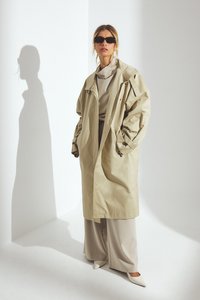 Bežs trenčkot ar pārmērīgiem piedurknēm, uzslāņots virs gaiši pelēka tērpa. Melni saulesbrilles, baltas norādītājus apavi, un redzama iemetamā ēna.