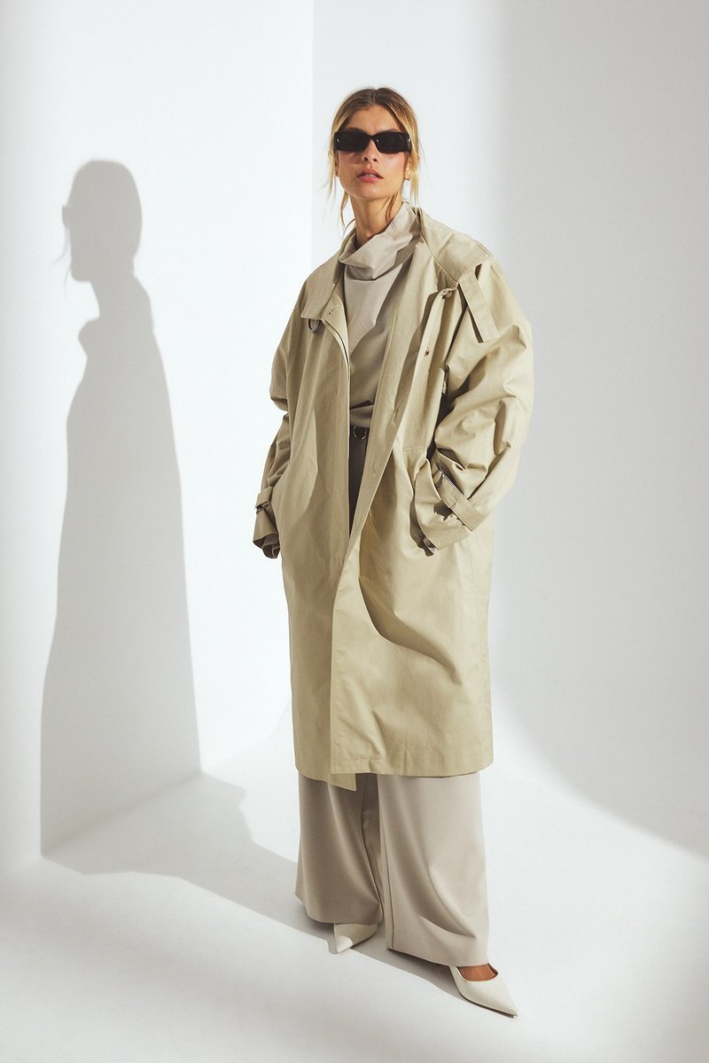 Bežs trenčkot ar pārmērīgiem piedurknēm, uzslāņots virs gaiši pelēka tērpa. Melni saulesbrilles, baltas norādītājus apavi, un redzama iemetamā ēna.