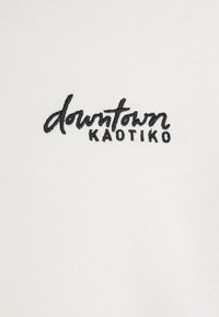 Tessuto bianco con testo ricamato nero "downtown KAOTIKO", caratterizzato da uno stile corsivo. Trama liscia e design minimalista.