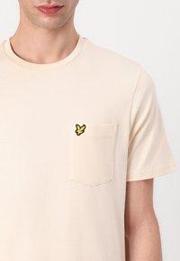 Beige bomull t-shirt med korta ärmar, rund halsringning och en bröstficka med en broderad fjärilsdesign i gult och svart.