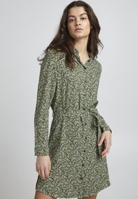 Abito button-up verde a fiori con maniche lunghe, vita legata e spacco laterale. Il tessuto presenta piccoli fiori bianchi e una consistenza morbida.