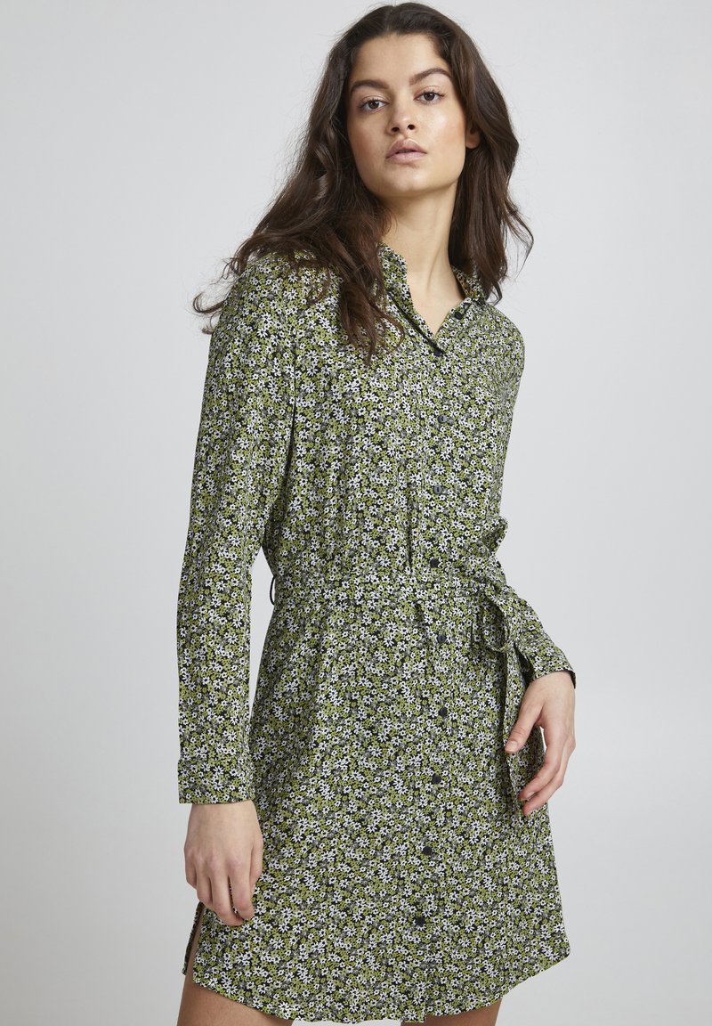 Abito button-up verde a fiori con maniche lunghe, vita legata e spacco laterale. Il tessuto presenta piccoli fiori bianchi e una consistenza morbida.
